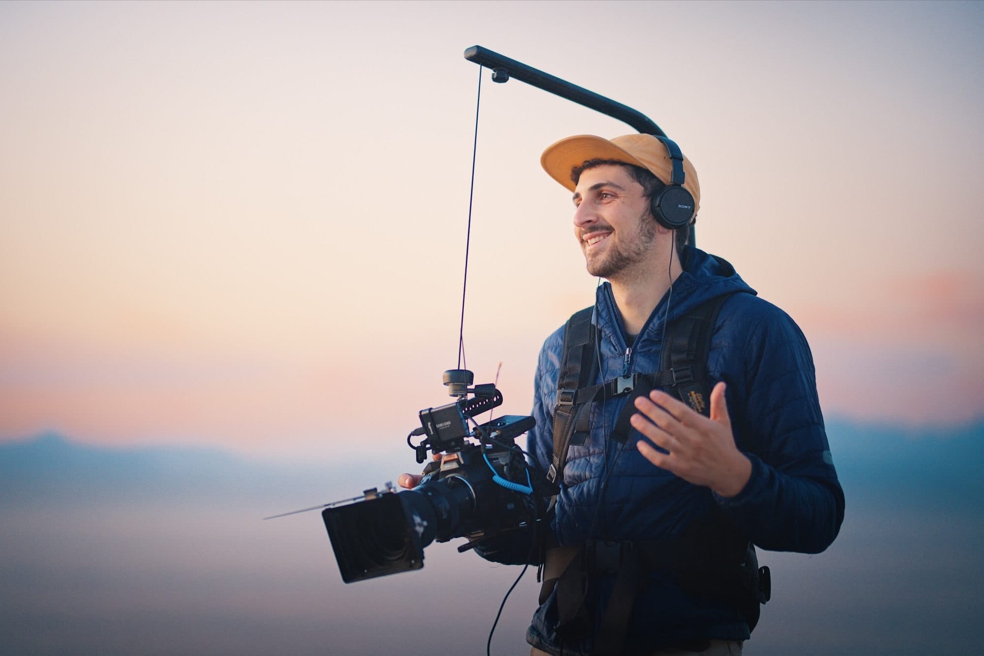 Benjamin Hazel smiling at sunrise, holding a cinema camera with a boom arm and wearing headphones